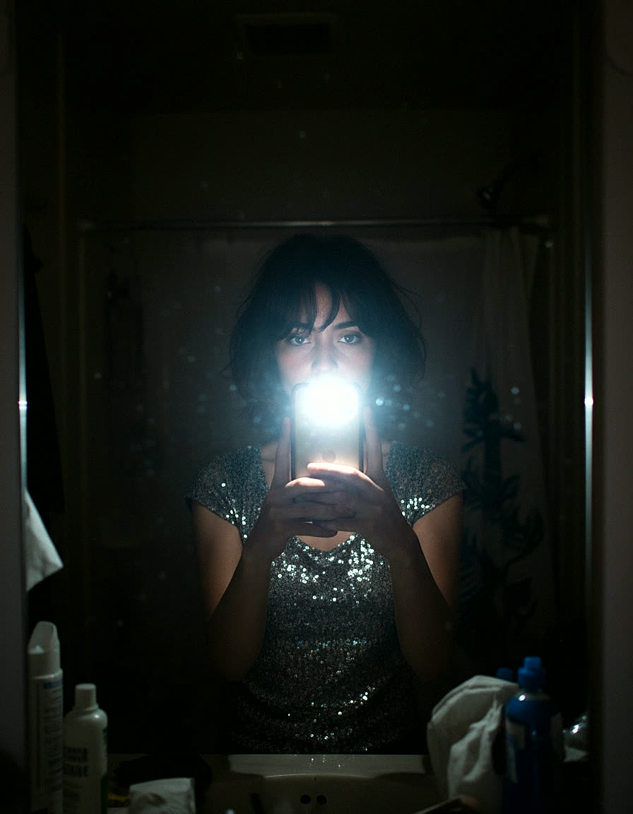 nadia — a candid phone-flash mirror selfie in a dark bathroom, sequin top, face lit by the flash